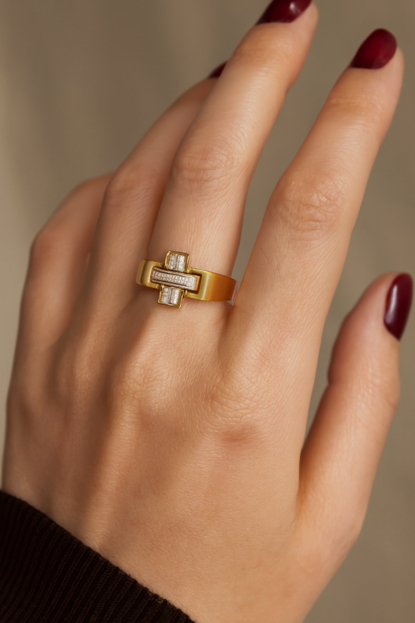 Gold ring with a diamond on a hand with maroon nail polish against a neutral background